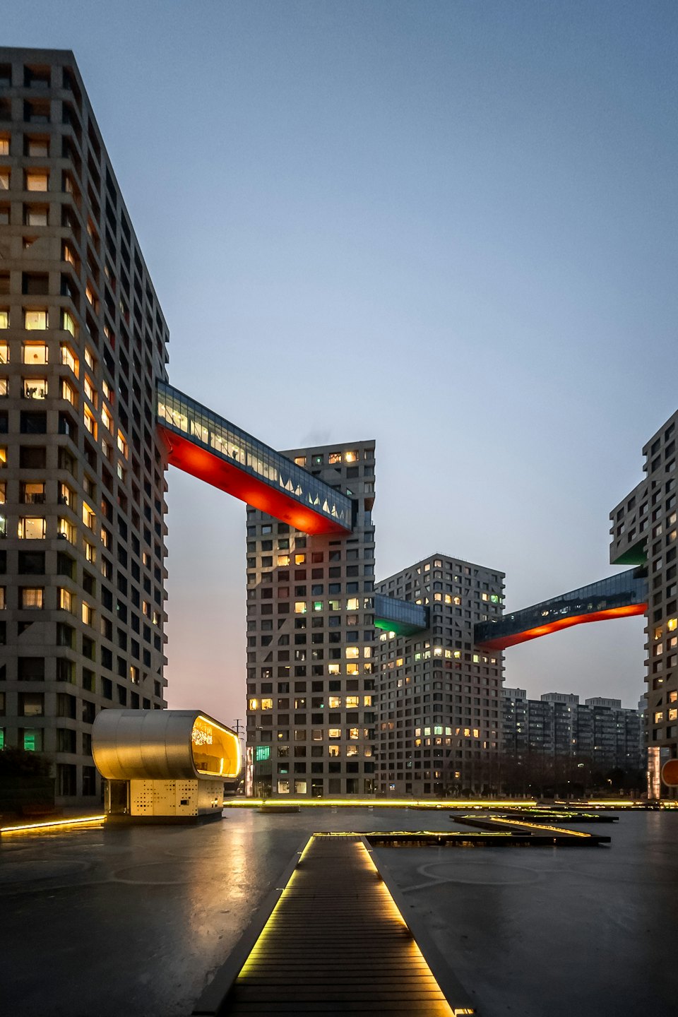 Modern concrete buildings connected by illuminated sky bridges at dusk.