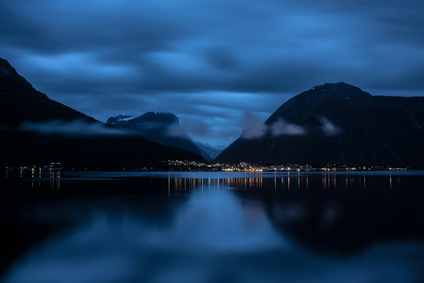 Small town lights reflect on water below dark mountain peaks.