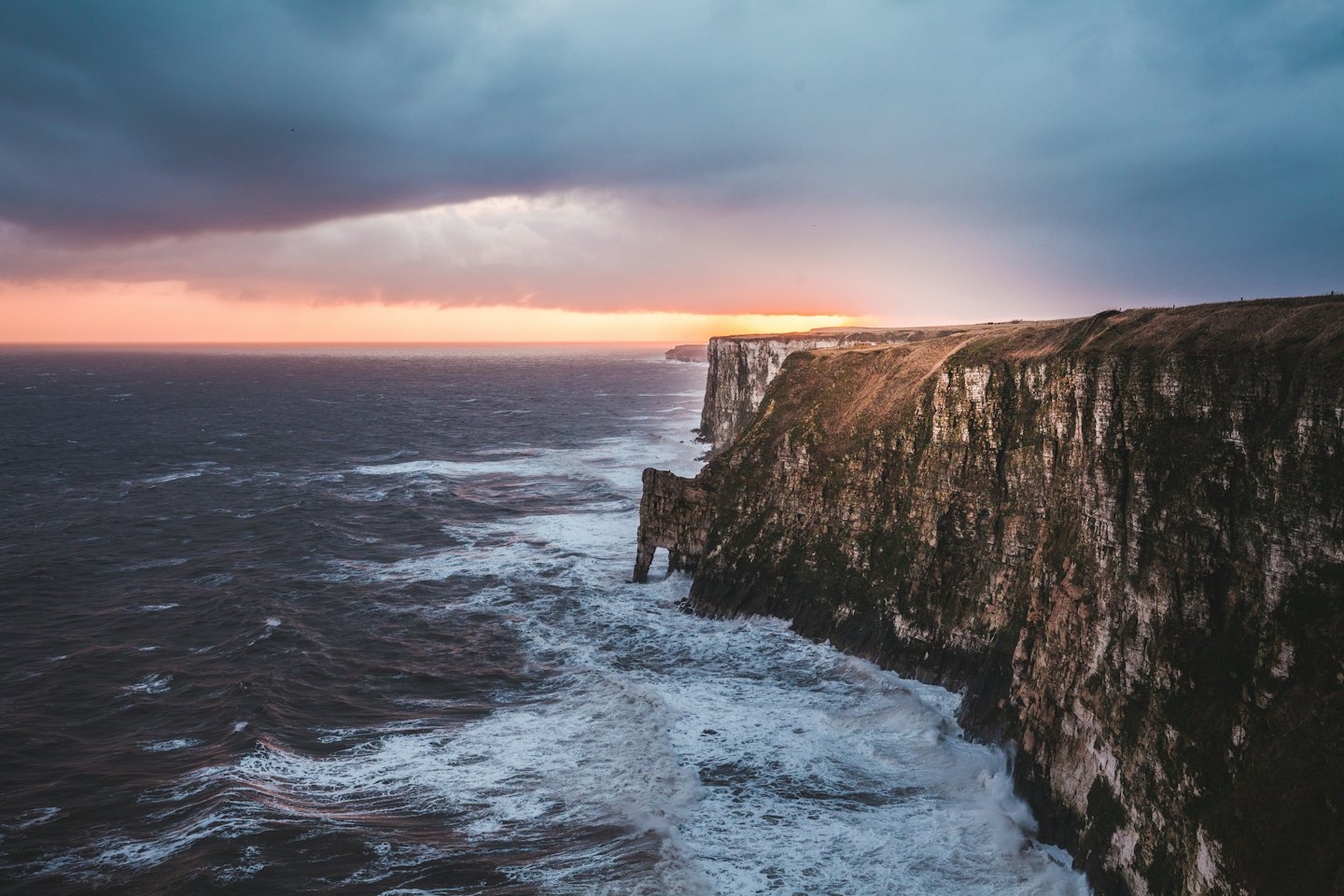 Waves crash against steep rugged cliffs under a stormy sky.