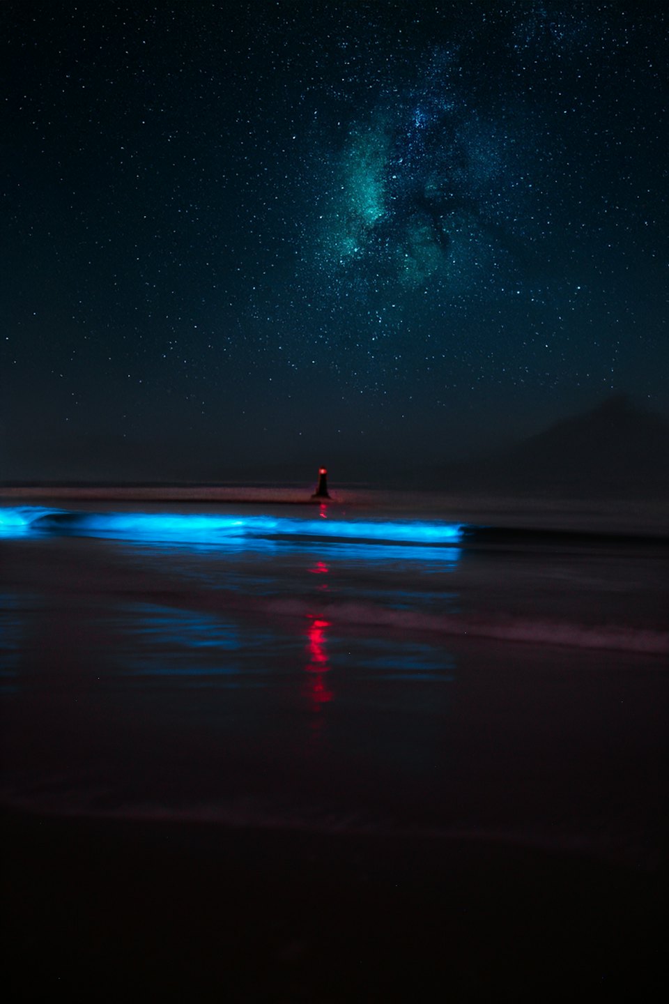 A lighthouse beam shines over bioluminescent waves and a starry sky.