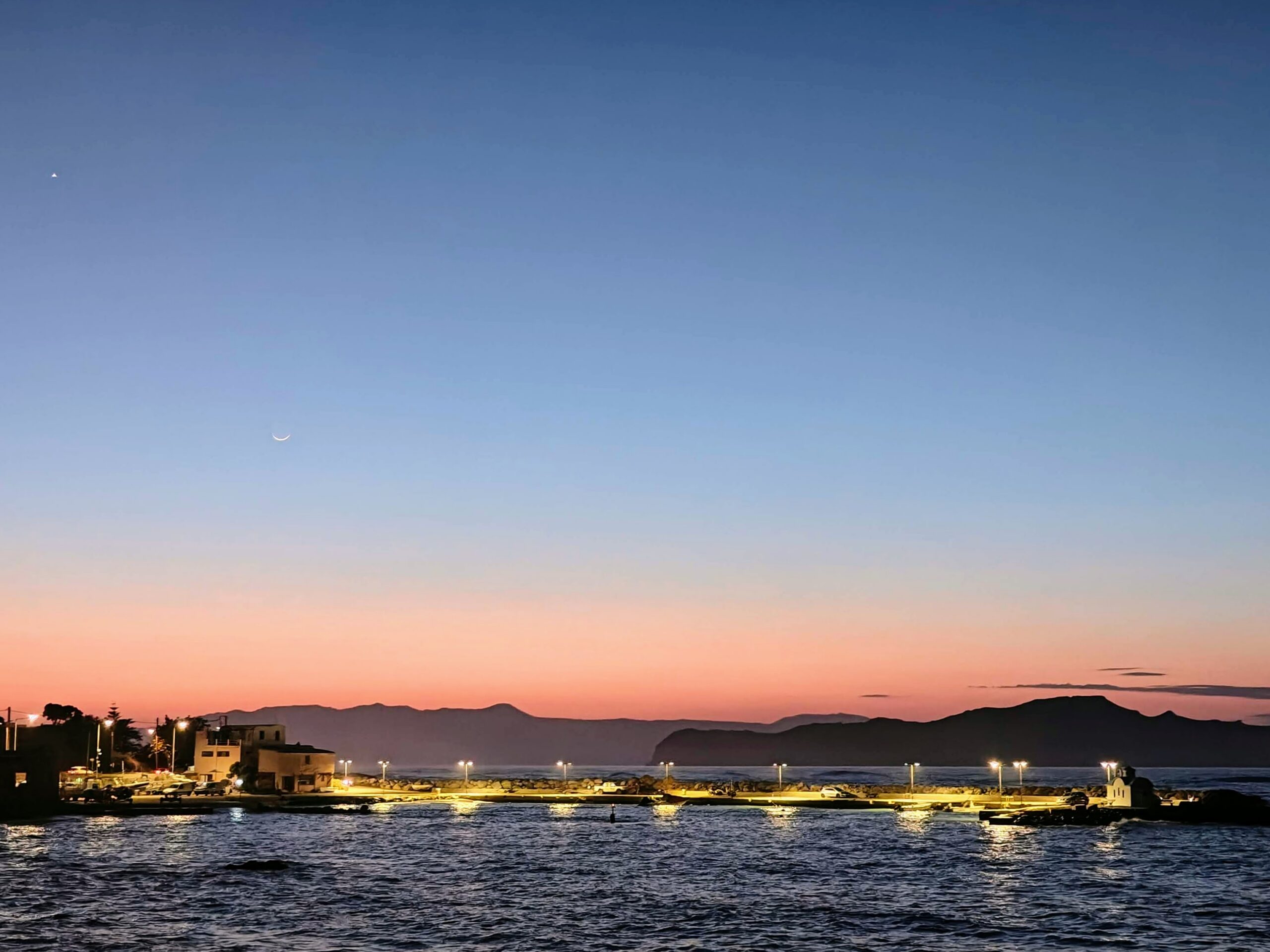 Dusk coastal scene in Budva: lit pier and buildings, rippling sea, silhouetted hills, crescent moon and a bright star.