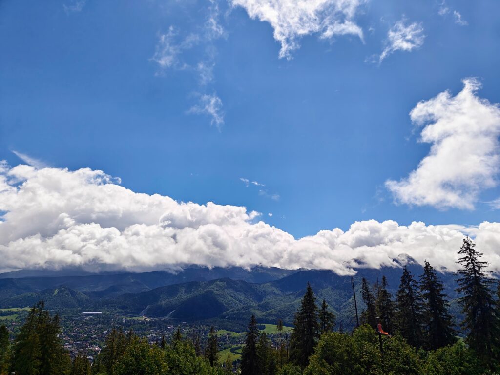 View of Zakopane valley with forested foreground, town below and mountains topped by low clouds under a blue sky