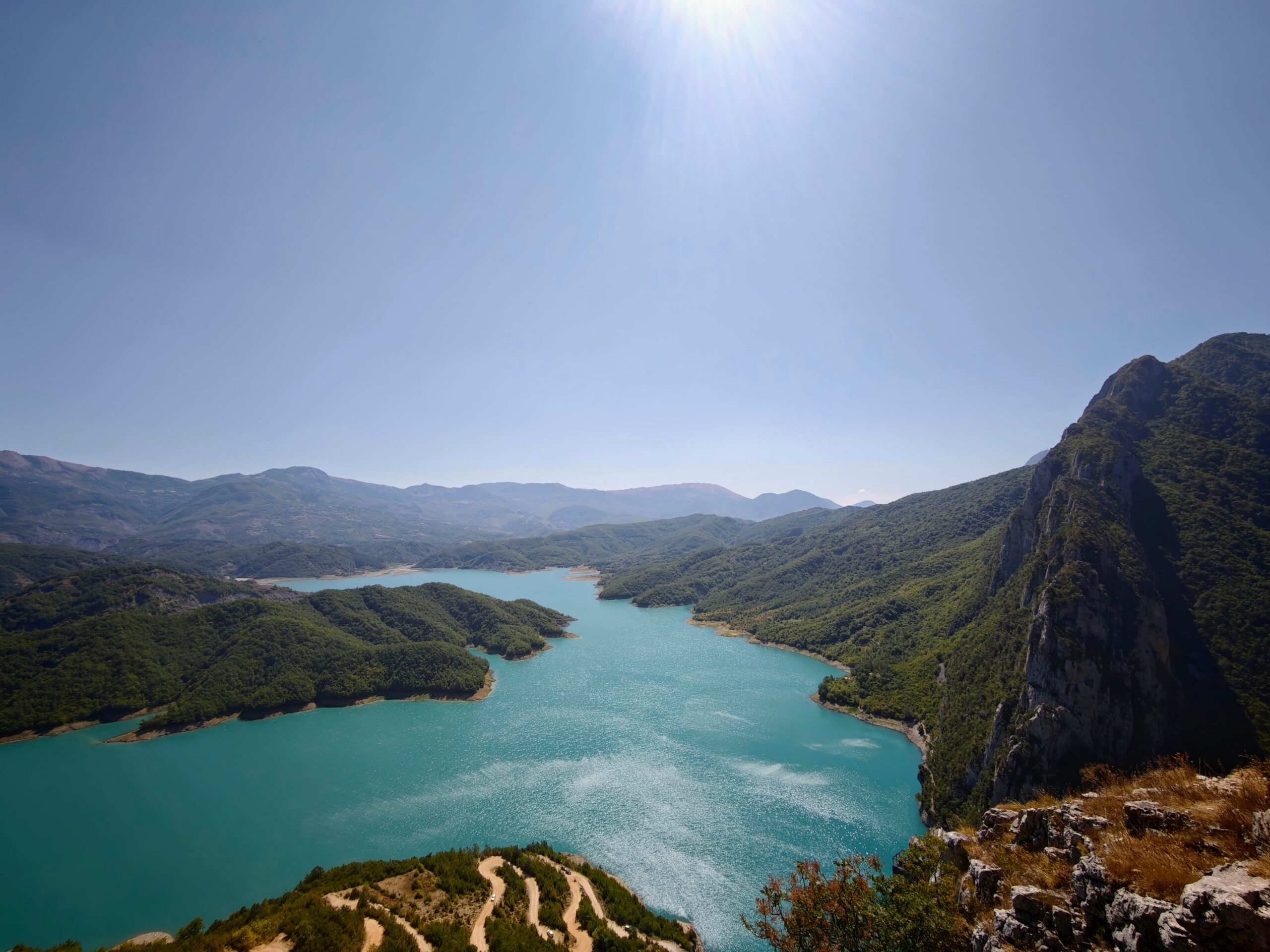 High viewpoint of turquoise reservoir winding through green hills and steep rocky cliffs under a clear sky.