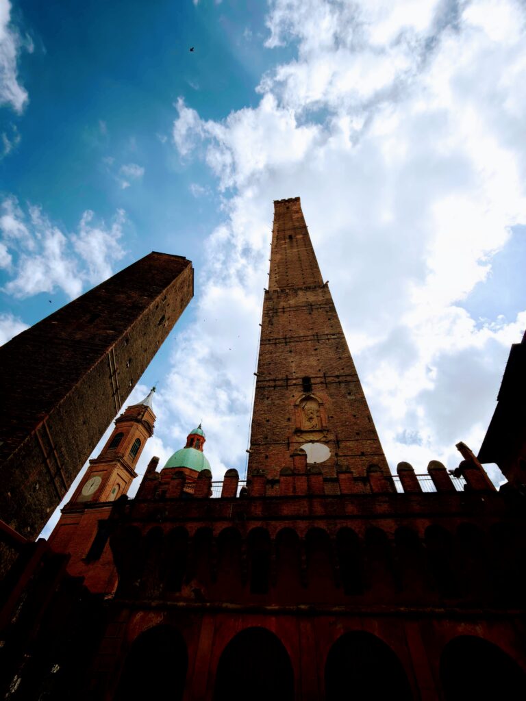 Low-angle view of two tall brick towers, domed church roofs and arched façade against a cloudy blue sky in Bologna.