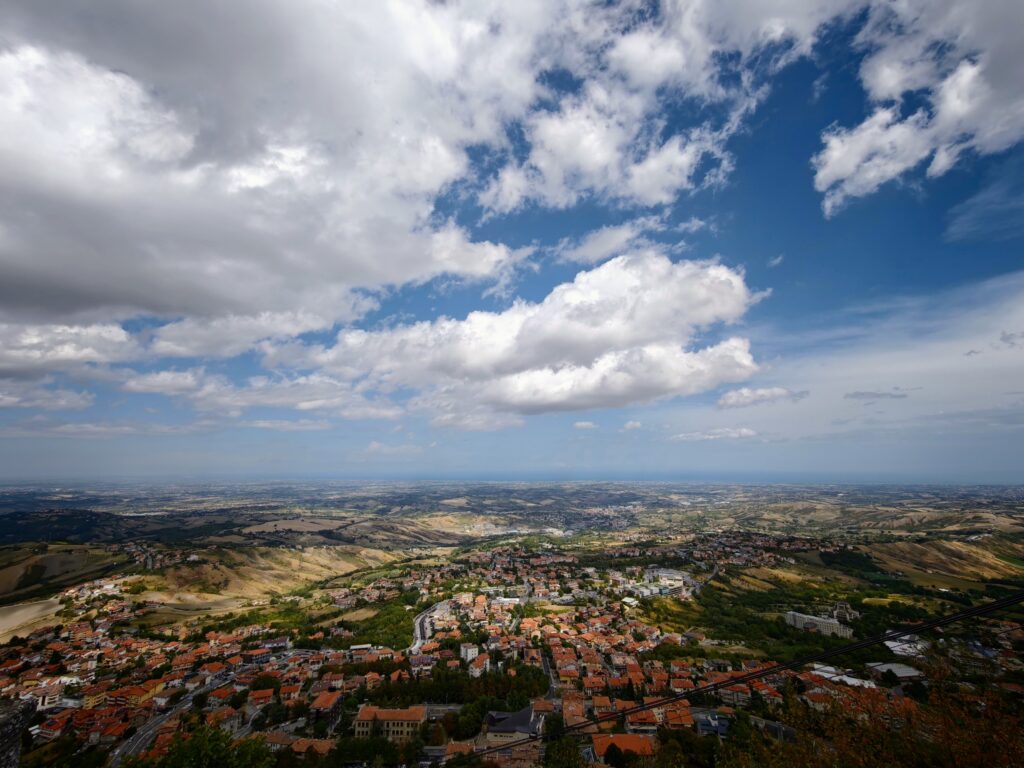 Panoramic view of San Marino town with terracotta rooftops, rolling fields and a vast sky filled with white clouds.