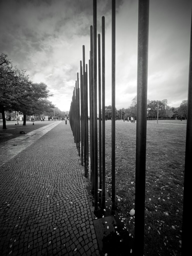 Black-and-white photo of tall vertical metal poles beside a cobbled path and grassy lawn in Berlin.