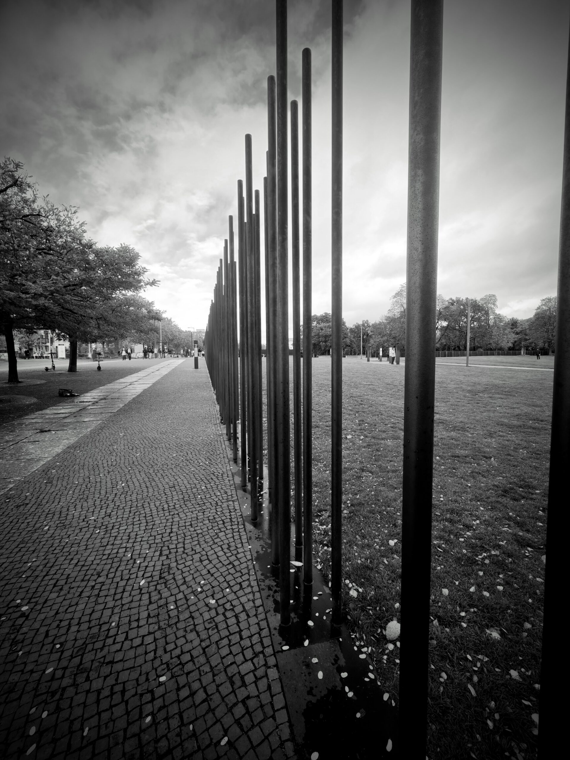 Black-and-white photo of tall vertical metal poles beside a cobbled path and grassy lawn in Berlin.