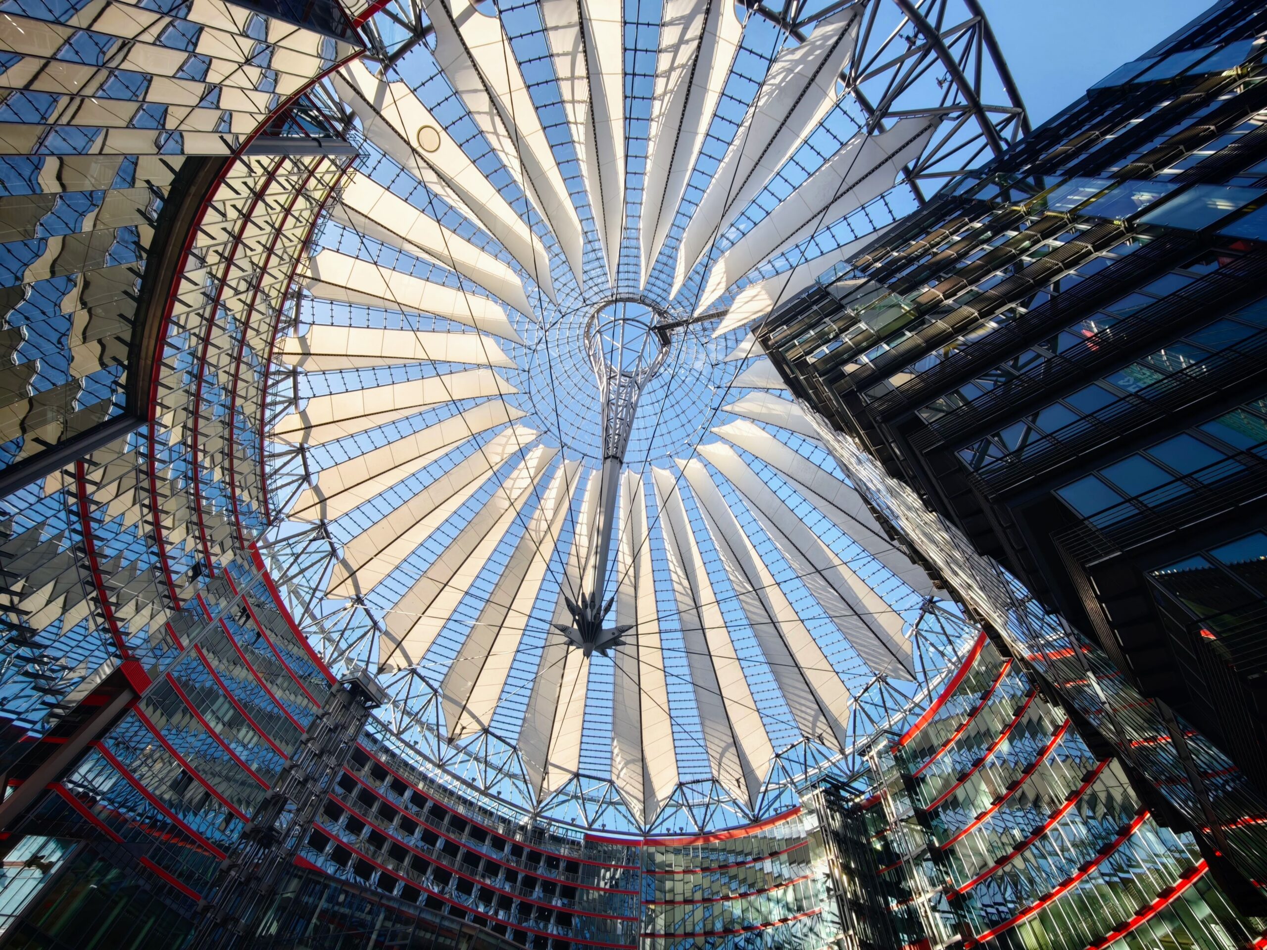 Upward view of a circular white fabric canopy with steel ribs and surrounding reflective glass buildings in a courtyard