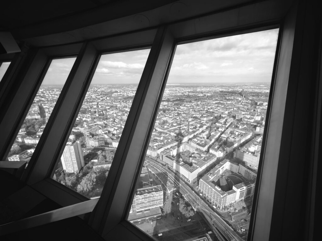 Black-and-white view of Berlin from high interior windows, showing rooftops, streets, and angled window frames.