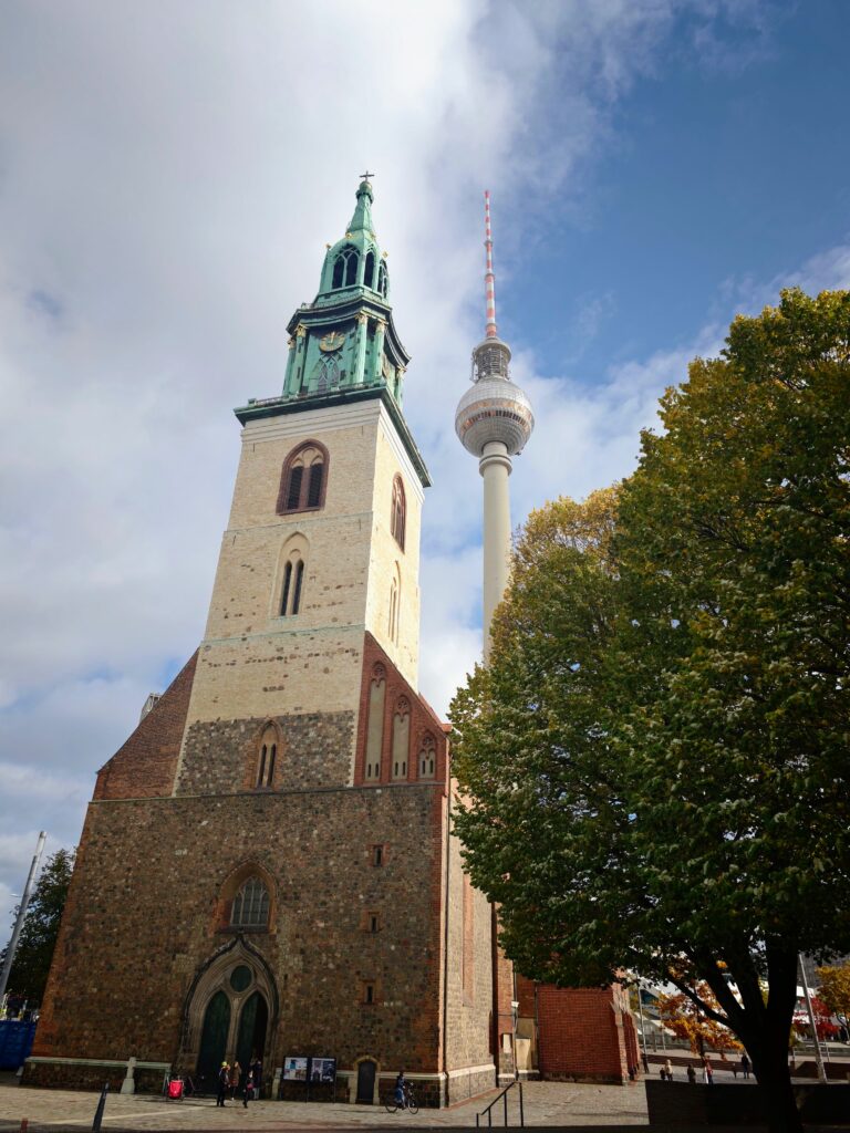 Low-angle view of a brick church tower, Berlin TV tower, a large tree, and a small plaza under a partly cloudy sky.